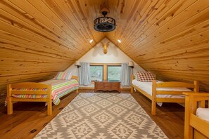 A cozy attic bedroom with wooden walls, a sloped ceiling, two beds, a dresser, and a large window. - Mtn Ski Retreat | Hot Tub + Fire Pit + Fireplace (Mars Hill)