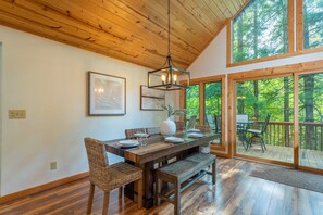 A dining area with a wooden table, chairs, and a bench. There are framed pictures on the wall, a hanging light fixture, and a view of the outdoors through large windows. - Mtn Ski Retreat | Hot Tub + Fire Pit + Fireplace (Mars Hill)