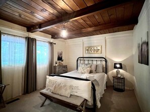 A bedroom with a wooden ceiling, a bed with white bedding, a bedside table with a lamp, and a window with curtains. - Mtn Ski Retreat | Hot Tub + Fire Pit + Fireplace (Mars Hill)