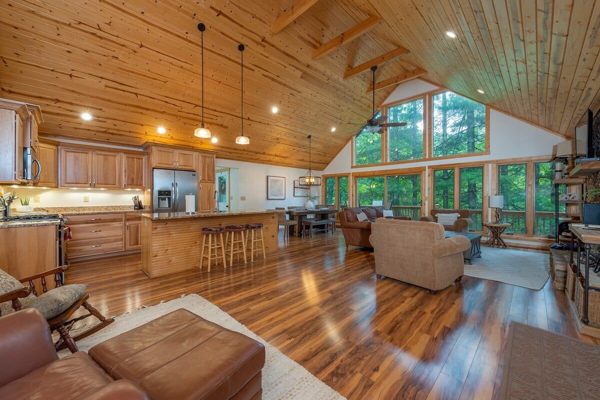 A spacious living area with a kitchen, wooden ceiling, large windows, and a cozy seating arrangement.