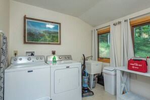 A laundry room with a washing machine, a sink, a window with curtains, and a red first aid kit on a shelf. - Mtn Ski Retreat | Hot Tub + Fire Pit + Fireplace (Mars Hill)
