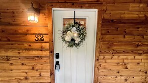 Eine Holztür mit weißem Klingelknopf, einer Hausnummer '32' und einem Blumengesteck.