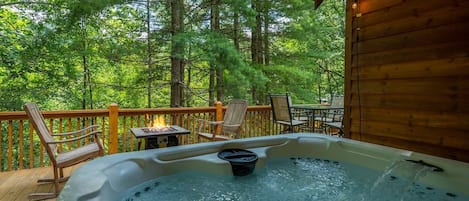 A hot tub on a wooden deck with chairs and a table, surrounded by a forest.