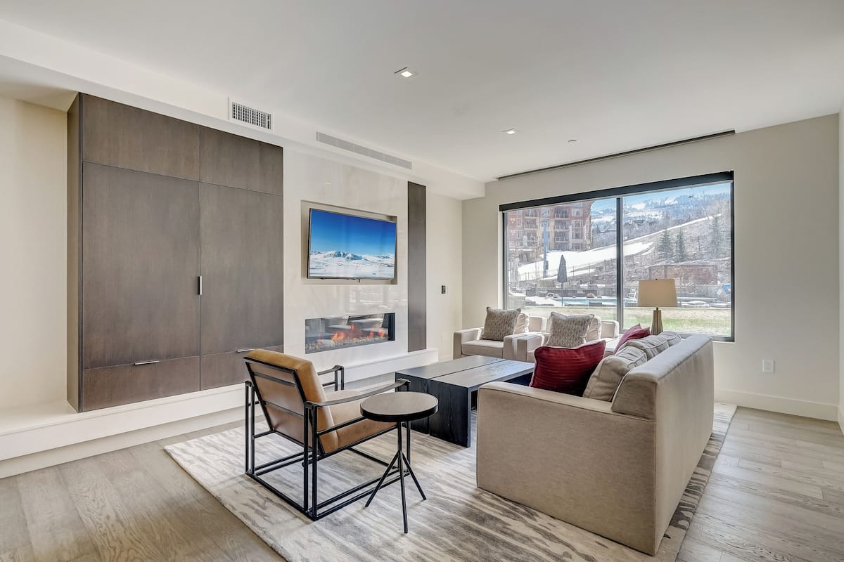 A modern living room with a fireplace, large windows, and a view of a snowy landscape.