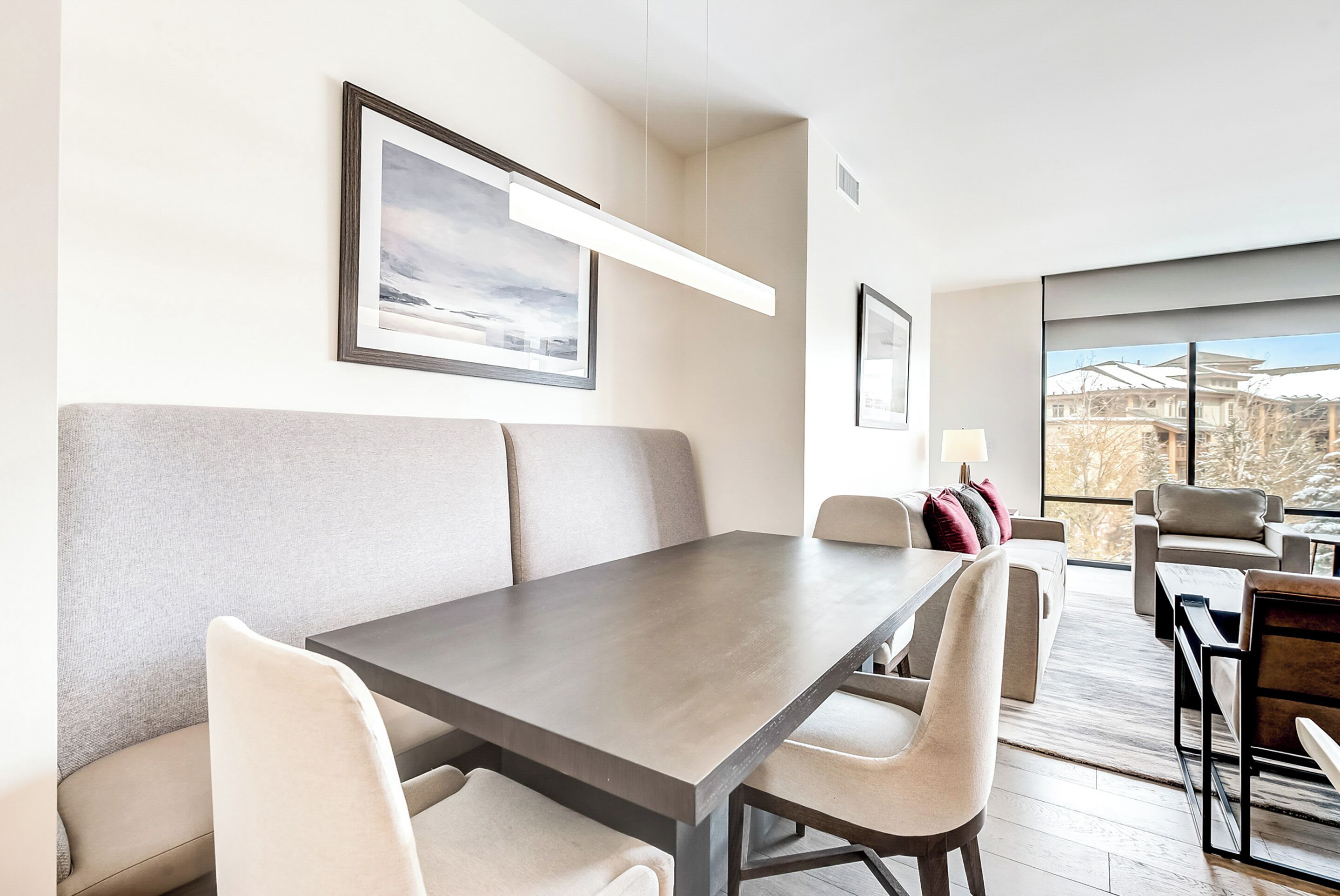A modern dining area with a long table and cushioned seating. There are framed pictures on the walls, and a large window offers a view of the outside.