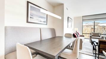 A modern dining area with a long table and cushioned seating. There are framed pictures on the walls, and a large window offers a view of the outside.