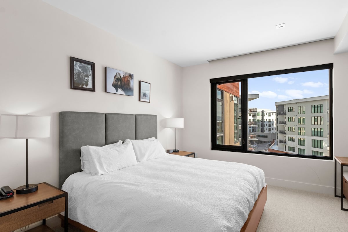 A modern hotel room with a large bed, two bedside tables, a nightstand, and a large window offering a city view.