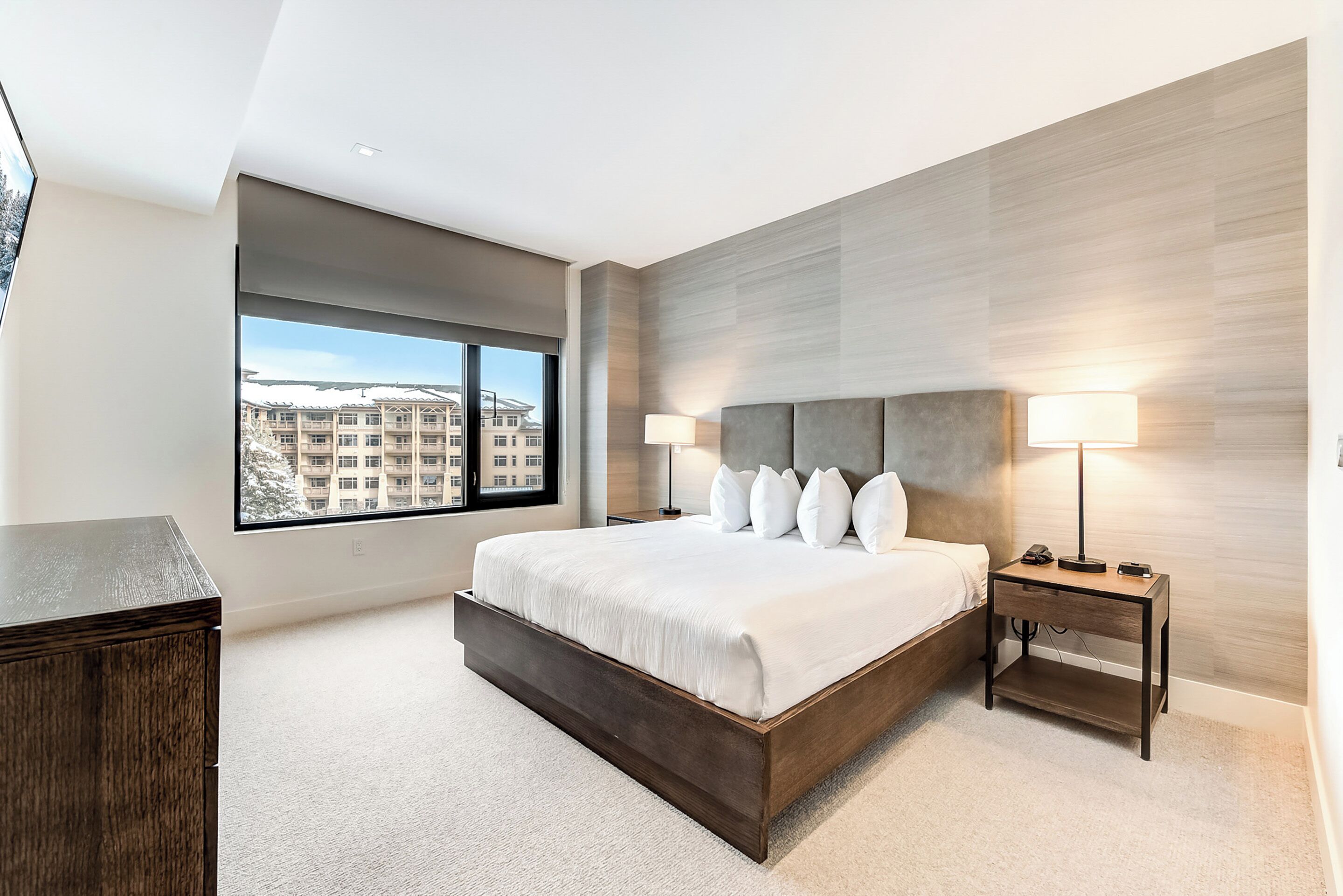 Habitación de hotel moderna con una cama grande, una mesita de noche, un escritorio y vistas a un paisaje urbano a través de un ventanal amplio.