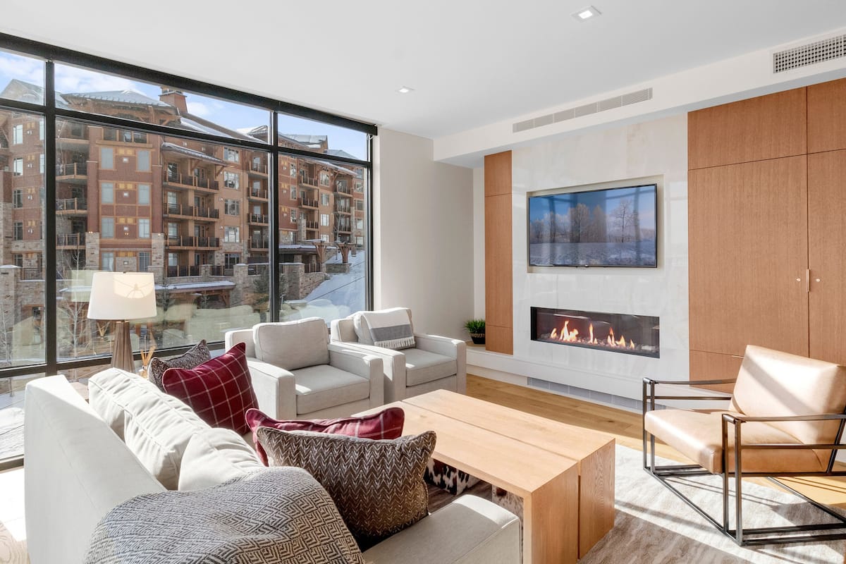 A modern living room with a fireplace, large windows, and a view of a residential area.