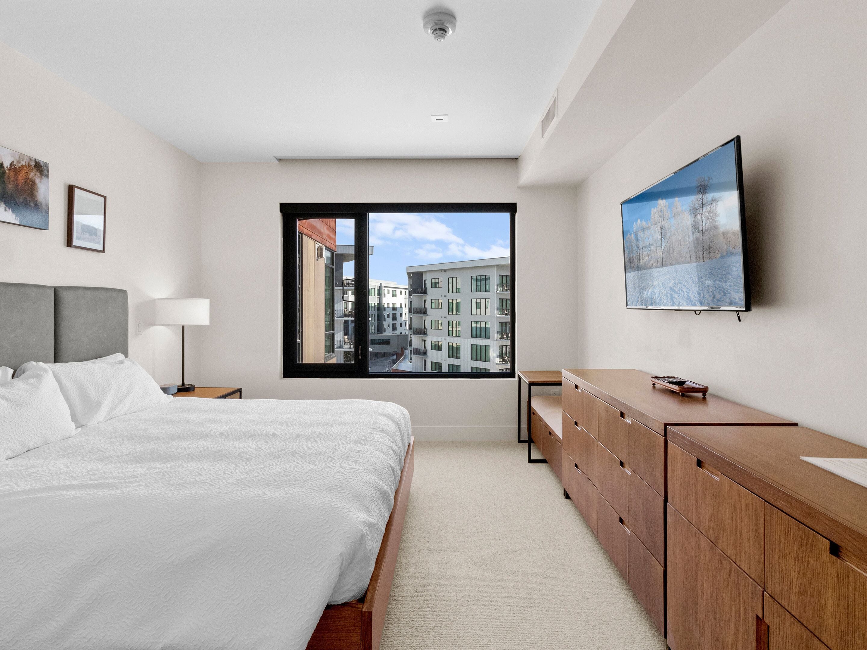 Ein modernes Schlafzimmer mit einem großen Bett, einer Holzkommode, einem Fernseher und einem Fenster mit Blick auf die Stadt.