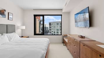 Ein modernes Schlafzimmer mit einem großen Bett, einer Holzkommode, einem Fernseher und einem Fenster mit Blick auf die Stadt.