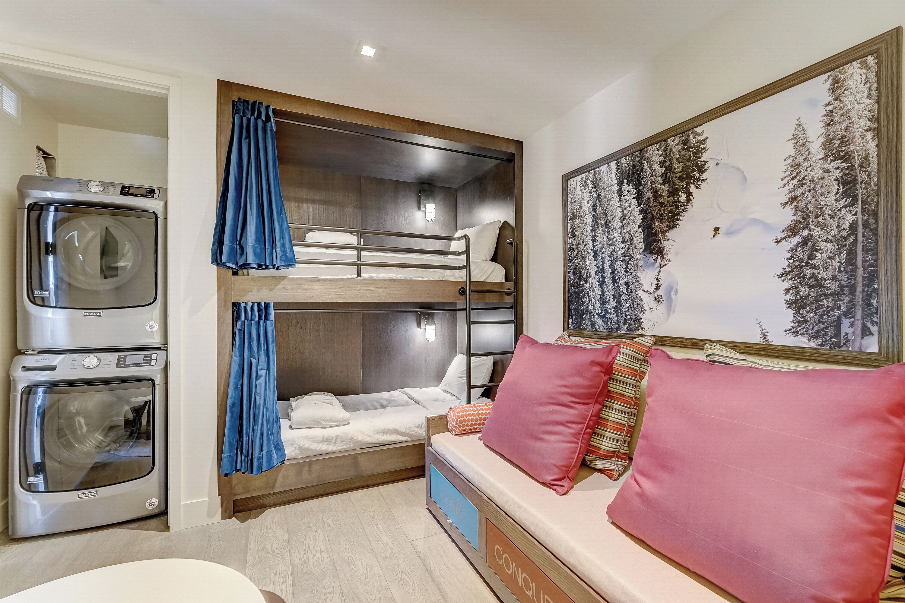 Una habitación de hotel con una cama litera, una lavadora, un sofá con un cojín rojo y un cuadro de un bosque nevado.