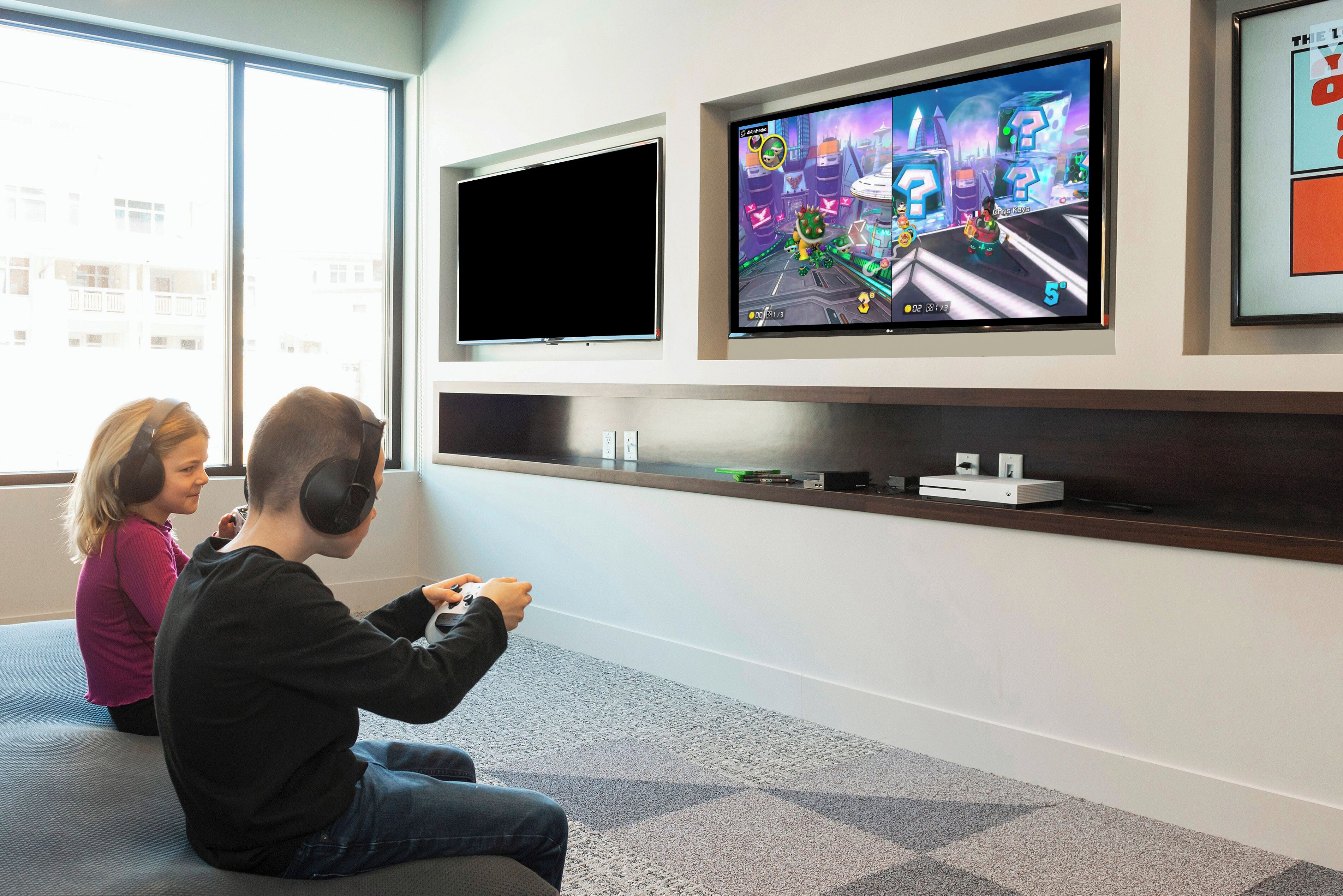 Two people playing a video game on a couch in a room with a large TV mounted on the wall.