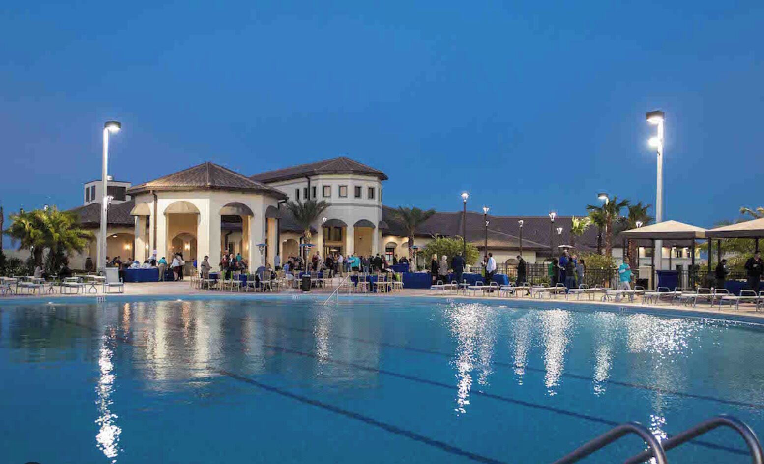 Une piscine à l’eau d’un bleu limpide, entourée d’un espace de détente couvert agrémenté de parasols, et un bâtiment au style architectural classique en arrière-plan.