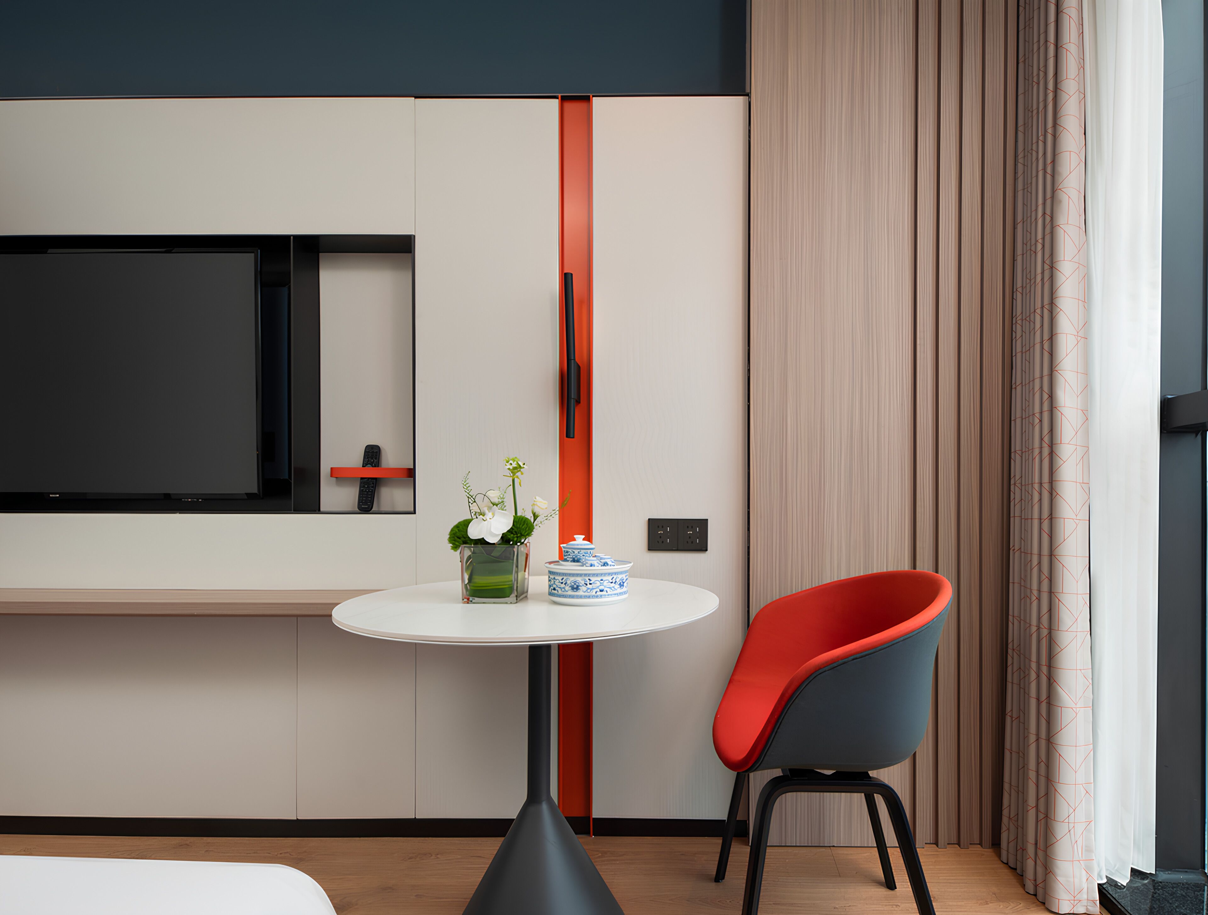 A modern hotel room with a flat-screen TV mounted on the wall, a small round table with a vase of flowers, a red chair, and a window with curtains.