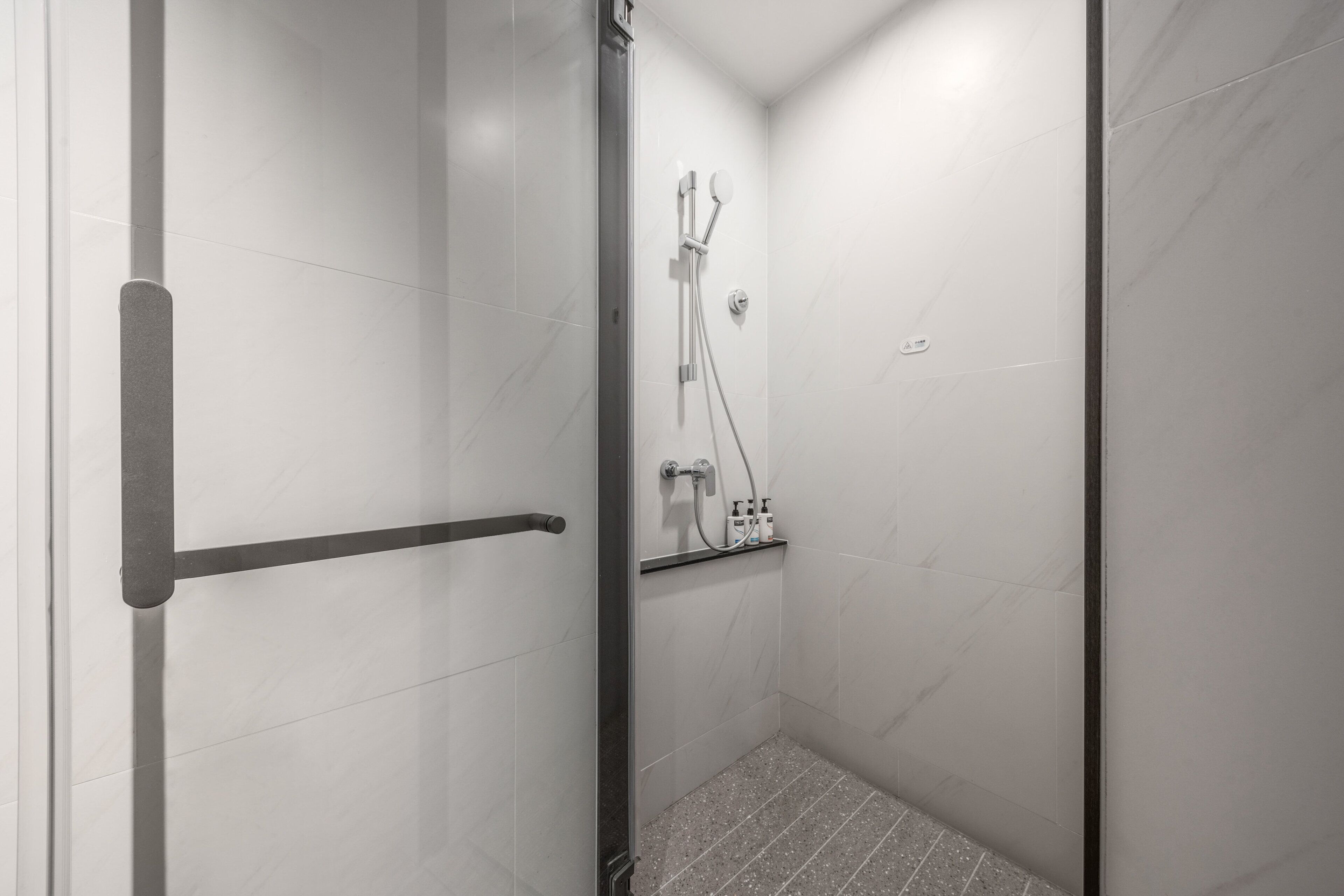 A modern shower with a glass door, a metal handle, and a shelf with toiletries.