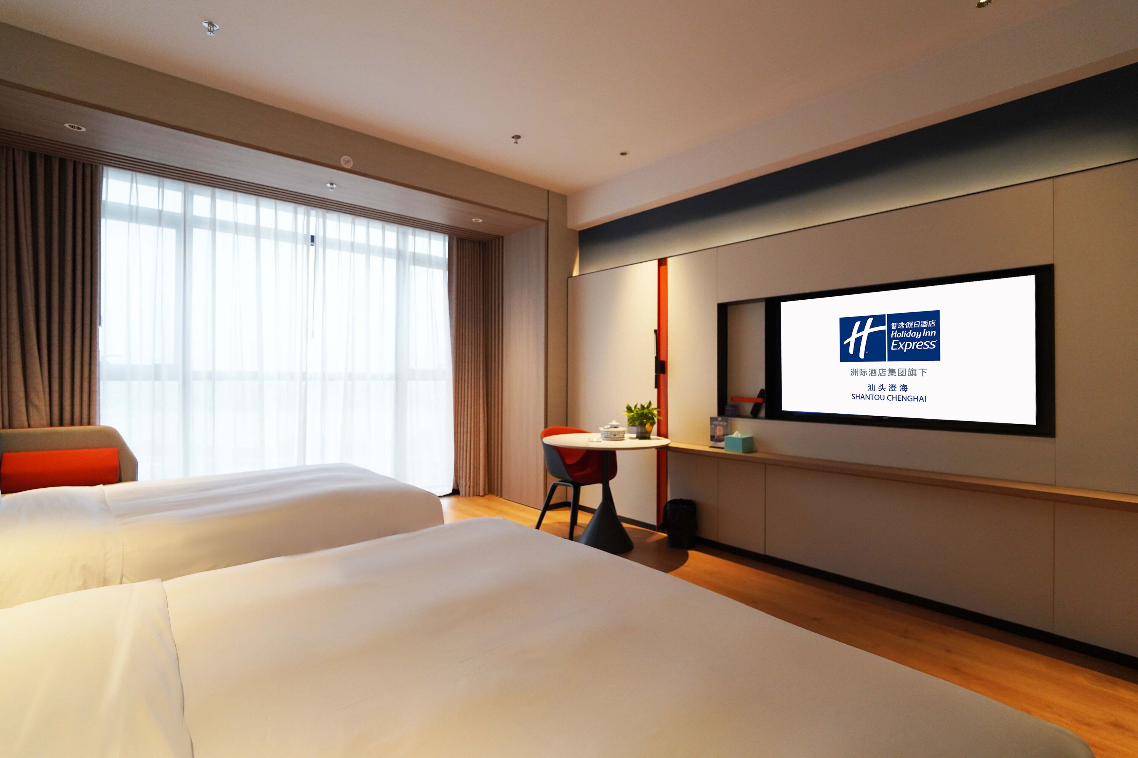 A hotel room with two beds, a desk, a TV screen displaying the hotel name, and a window with curtains.