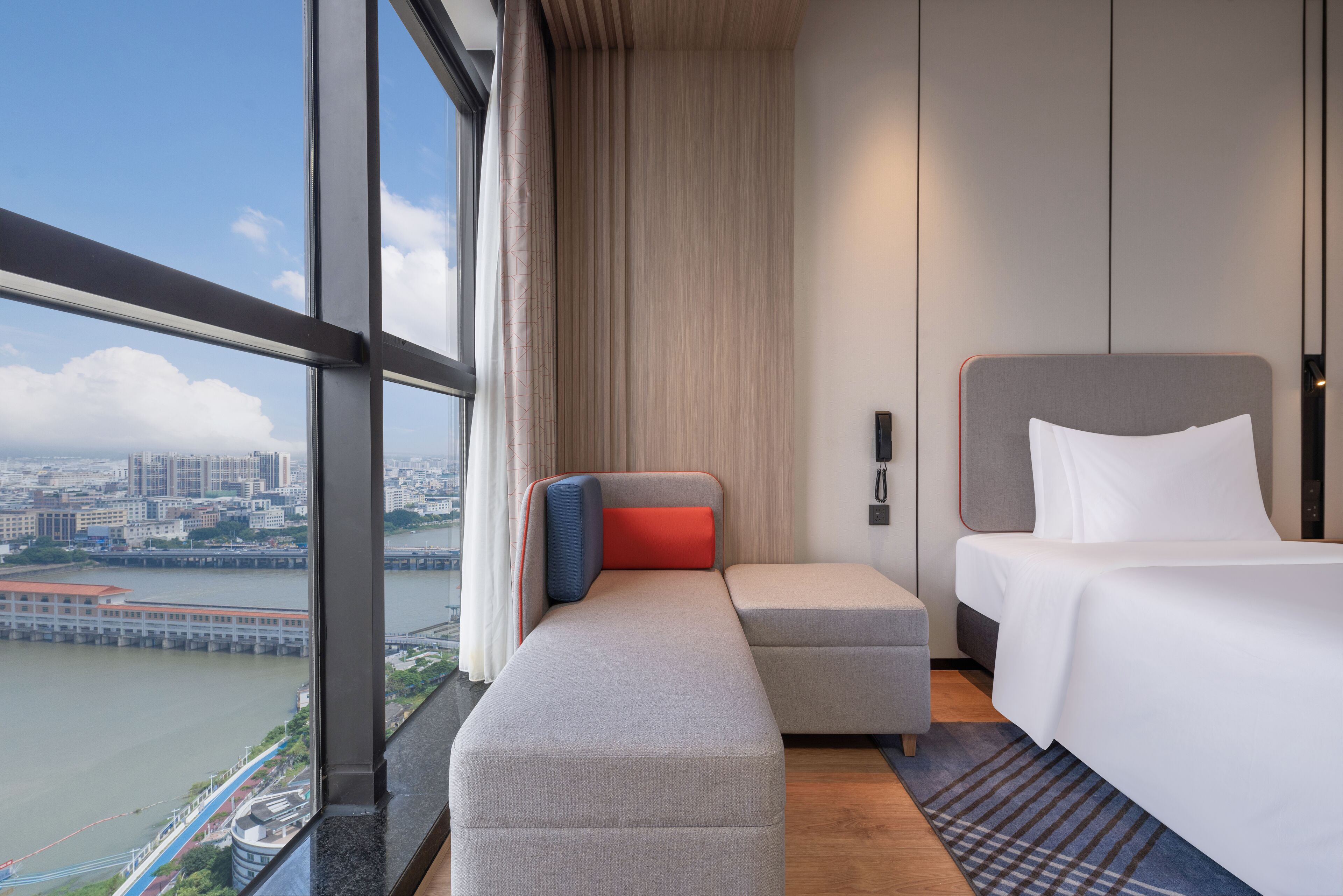 Una habitación de hotel moderna con un ventanal grande, una cama, un banco y vistas a un paisaje urbano y un río.