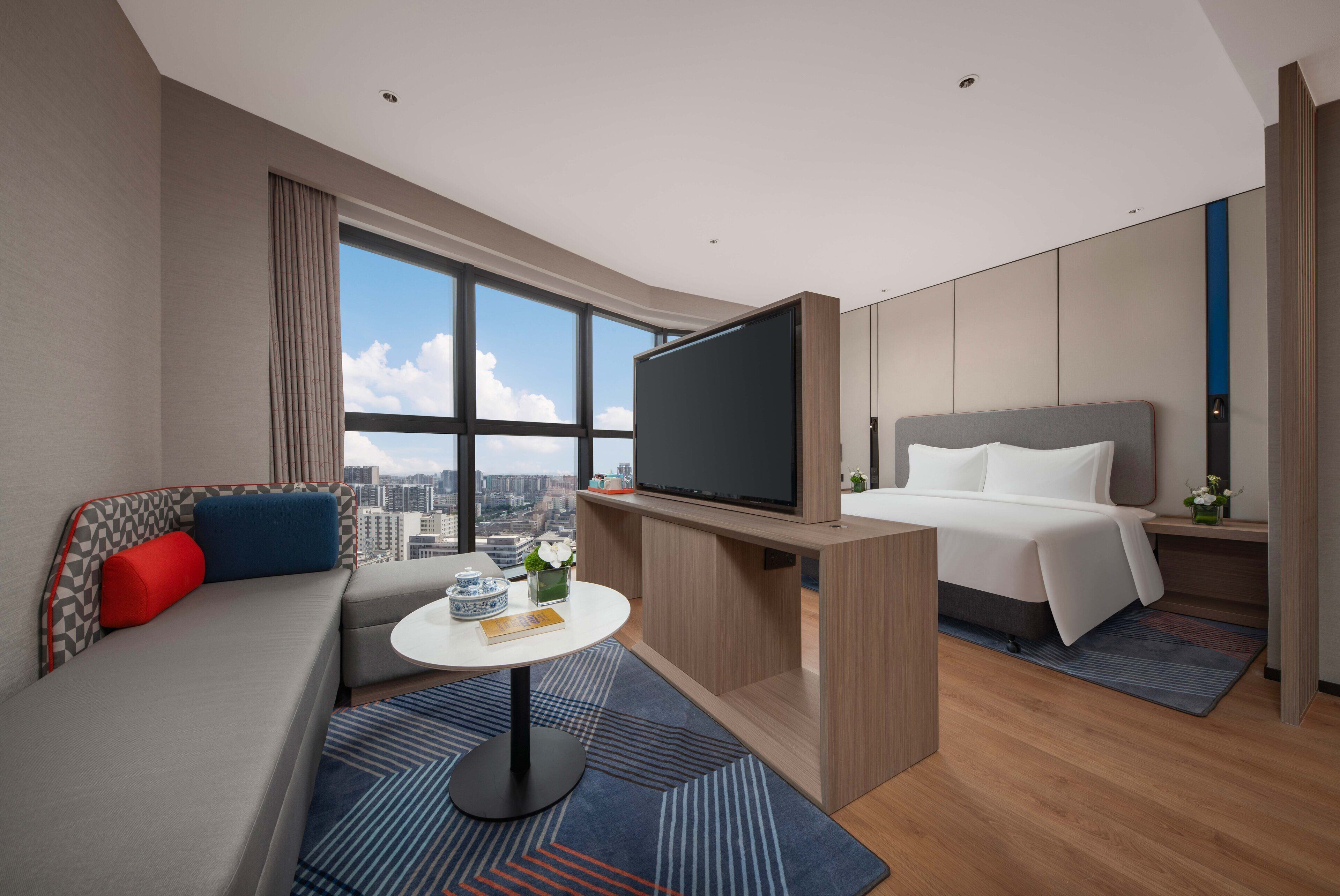 A modern hotel room with a sofa, a bed, a TV, a small table, and a city view.