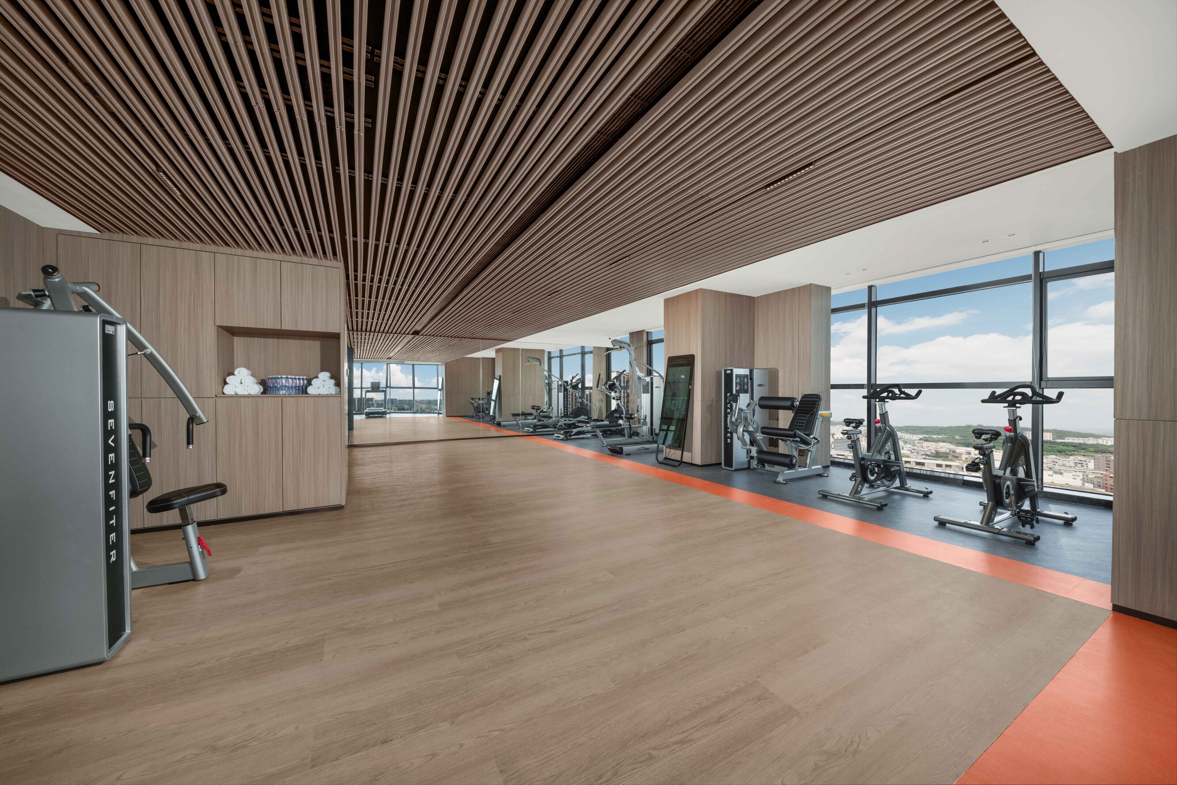Une salle de sport moderne dotée de planchers en bois, d’équipements d’exercice et de grandes fenêtres offrant une vue sur la ville.