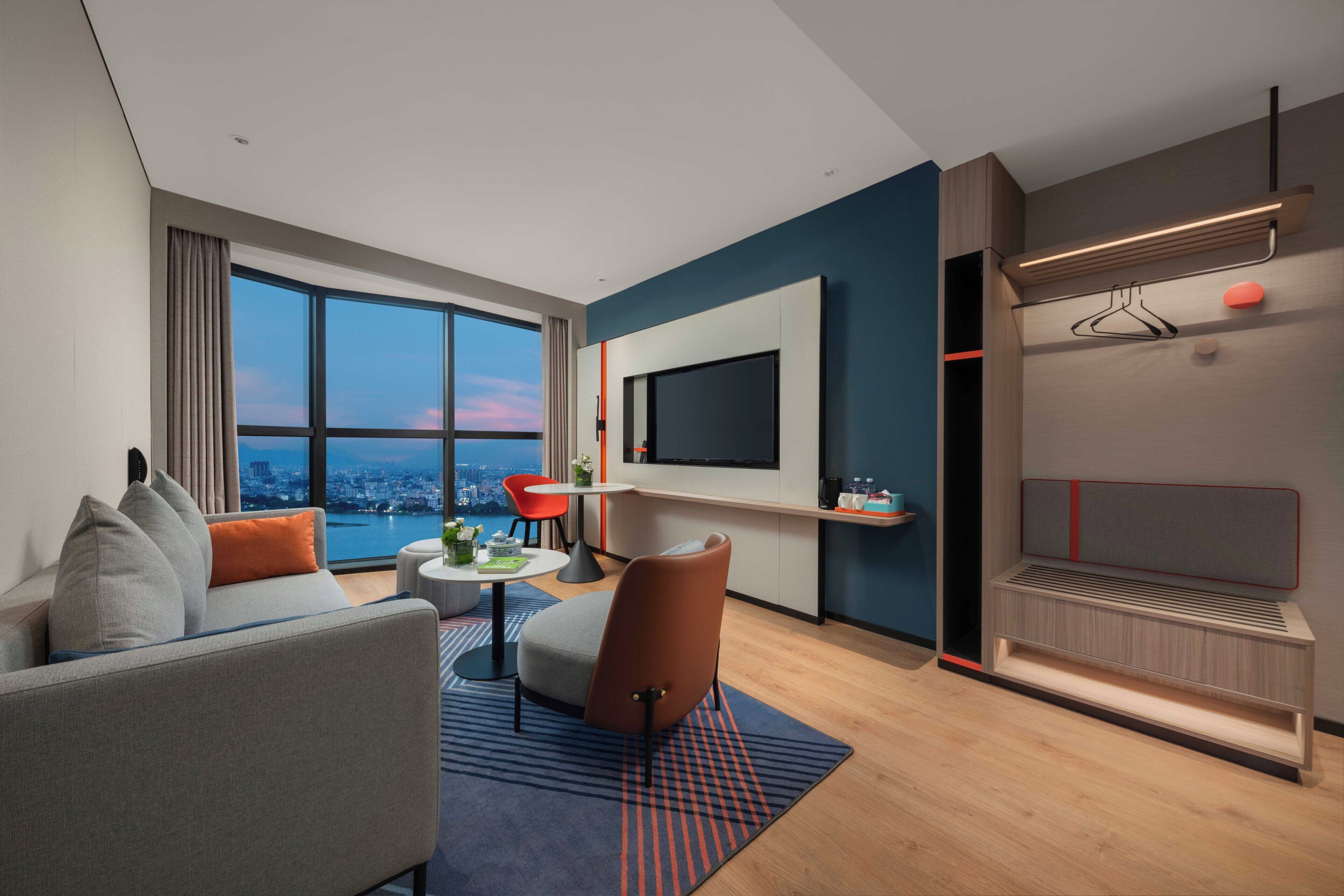 A modern hotel room with a large window, a sofa, a coffee table, a TV, and a built-in bed.
