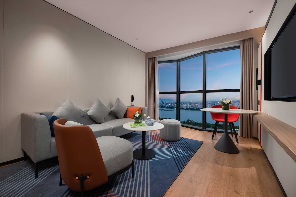 A modern hotel room with a sofa, a small round table, a red chair, and a large window offering a city view.