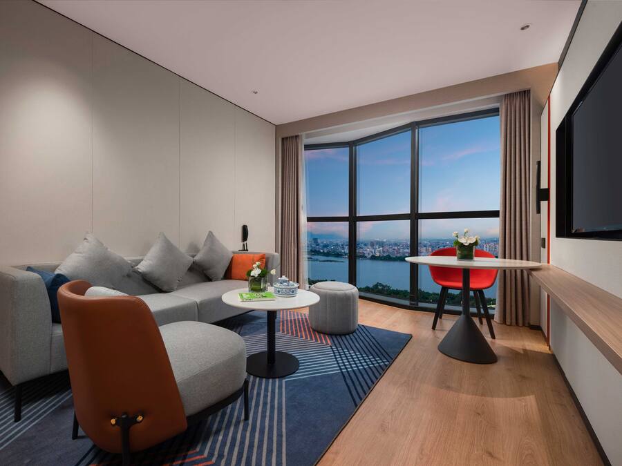 A modern hotel room with a sofa, a small round table, a red chair, and a large window offering a city view.