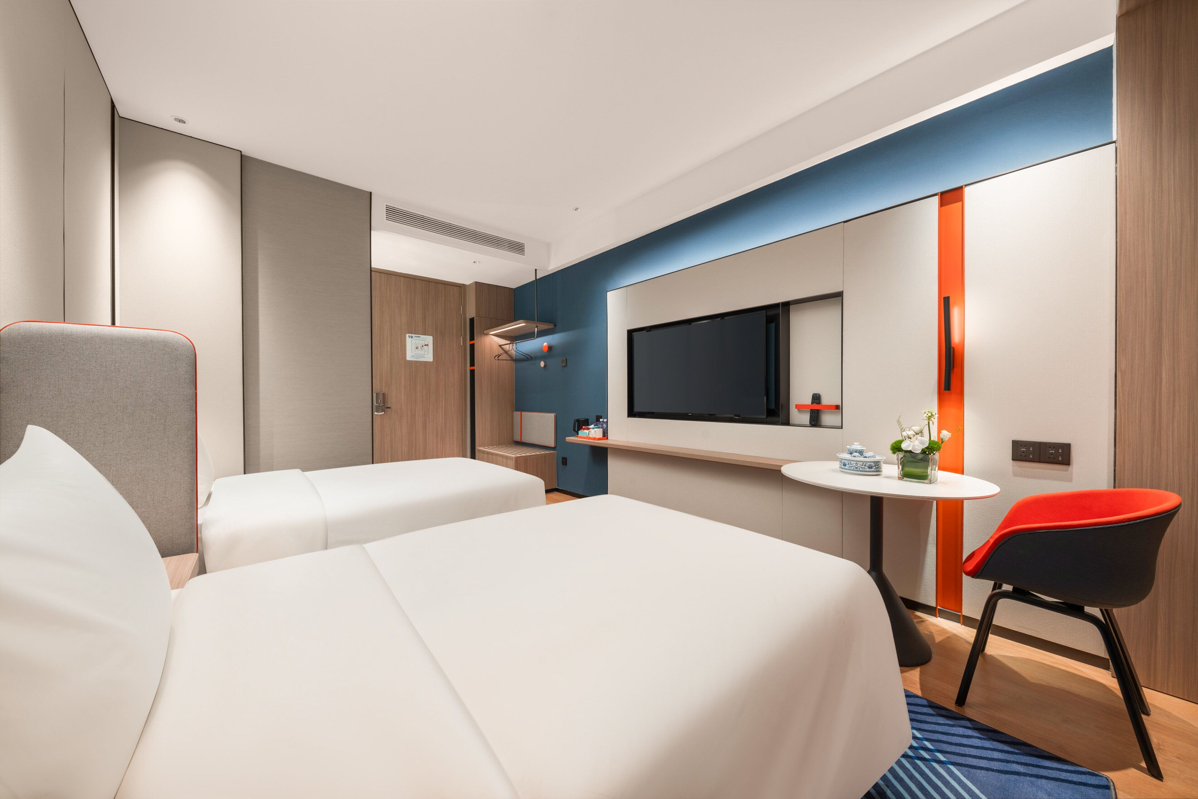 A modern hotel room with two beds, a TV, a small table, and a red chair.
