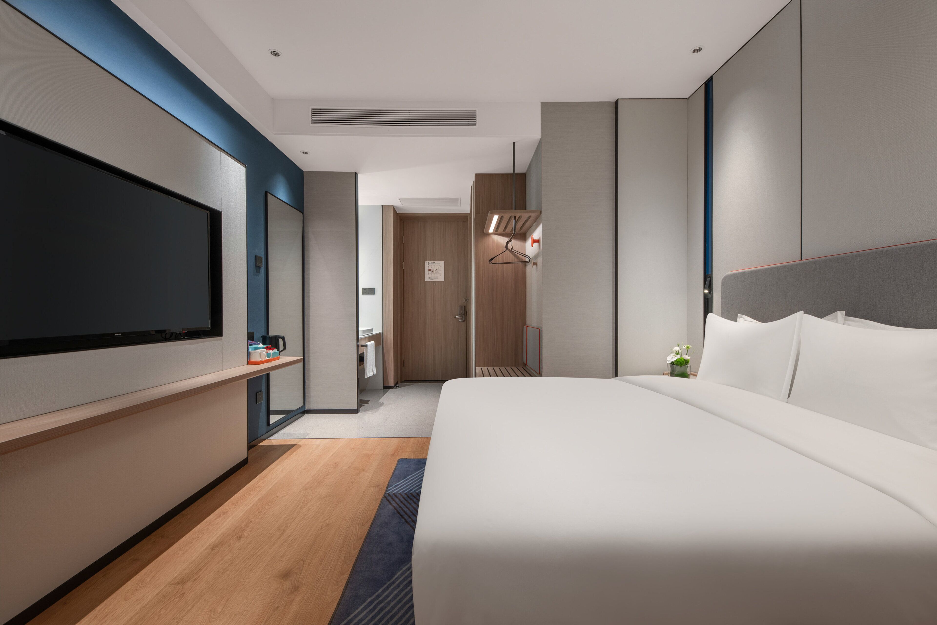 A modern hotel room with a large bed, a flat-screen TV, a wooden floor, and a bathroom visible in the background.