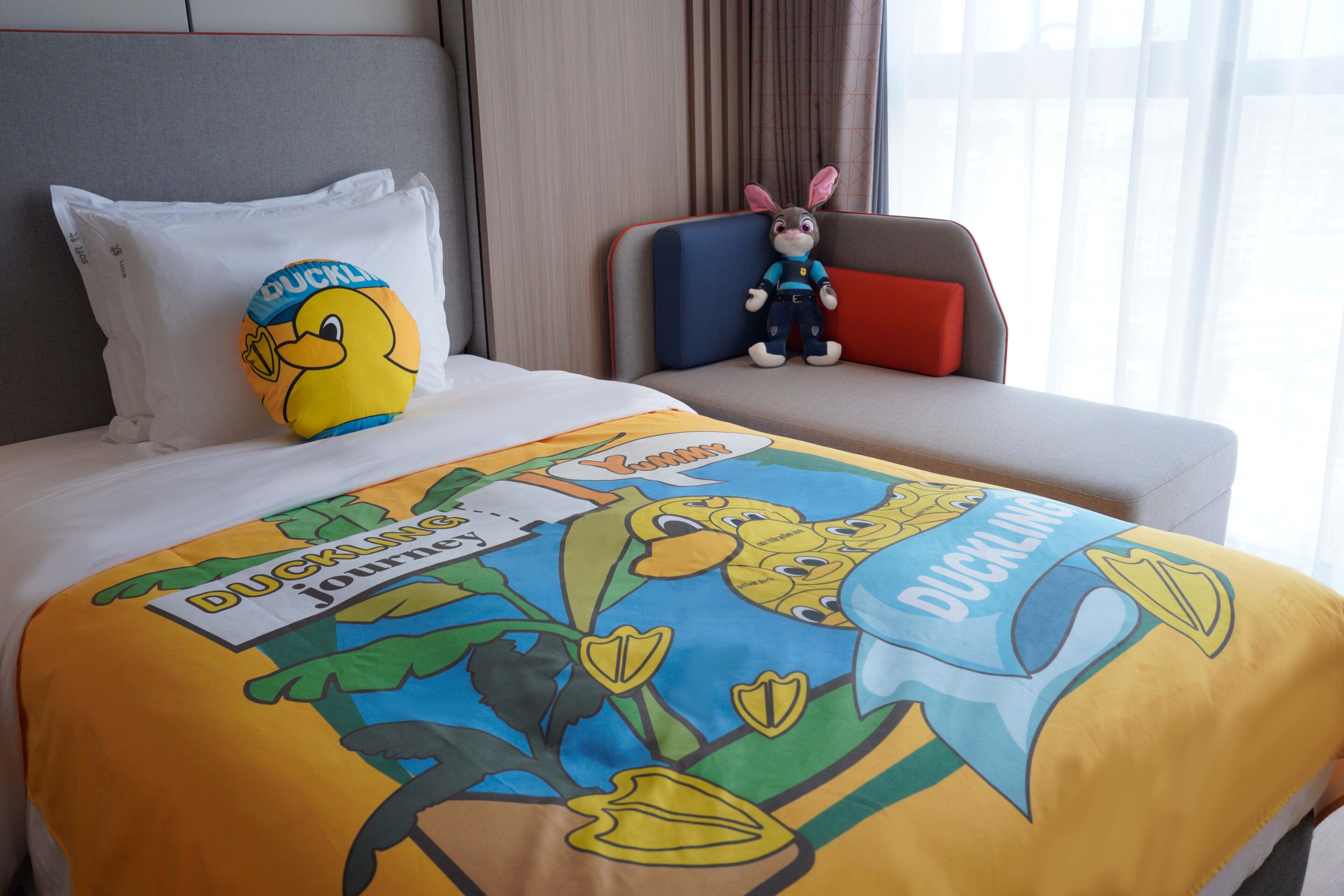 Ein Hotelzimmer mit einem Bett, das mit einer Enten-Motiv-Bettdecke, einem Kuscheltier und einem Kopfkissen mit dem Enten-Logo ausgestattet ist.