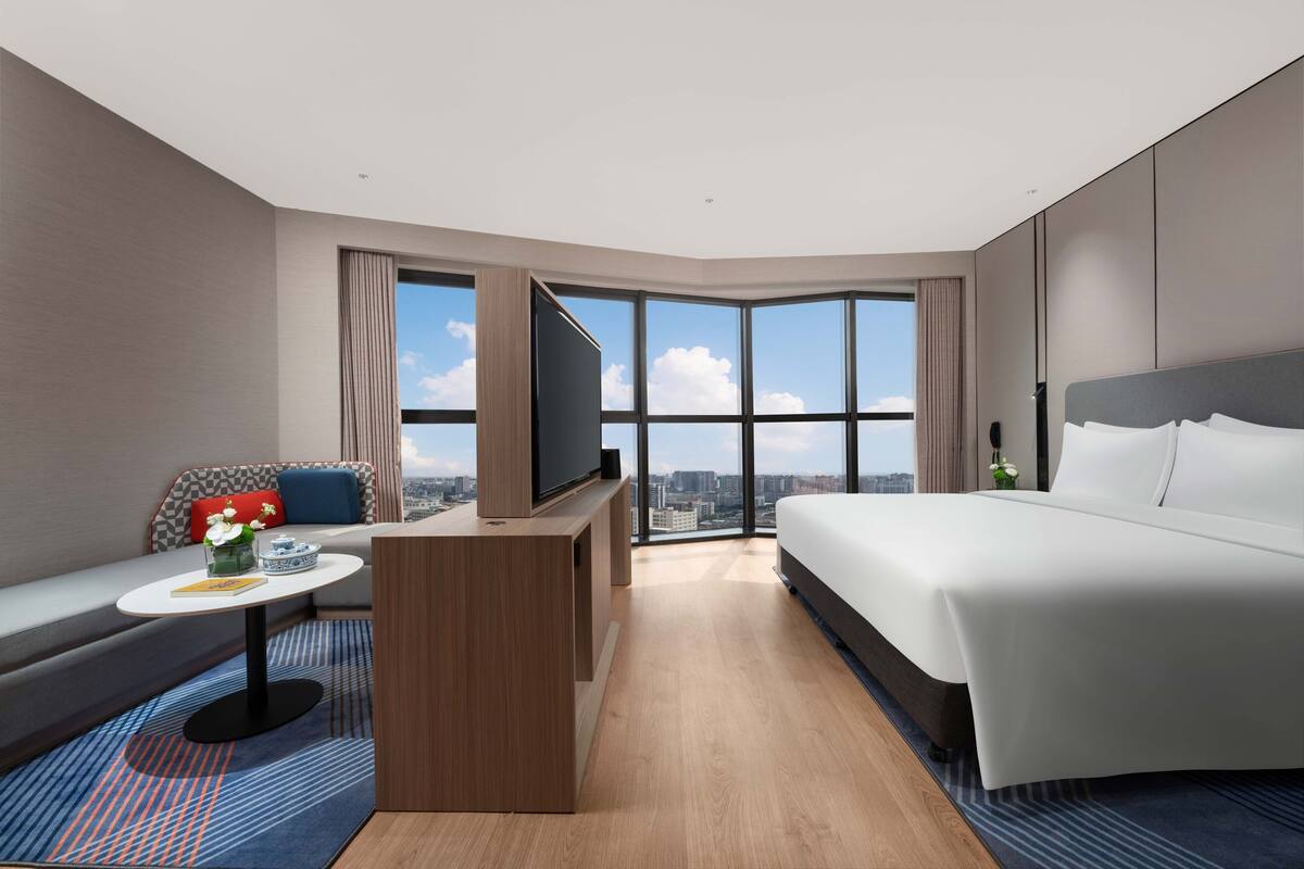 A modern hotel room with a large bed, a sofa, a TV on a wooden stand, and a city view through large windows.