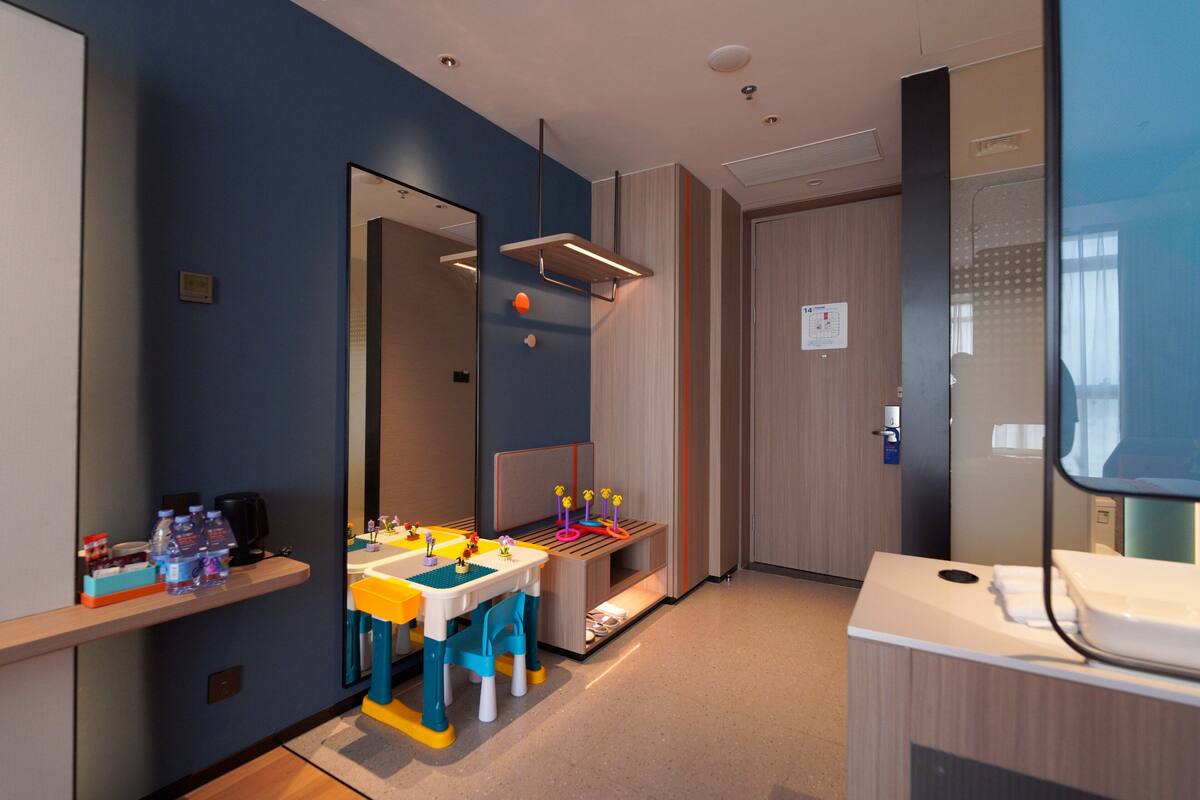A modern hotel room with a large mirror, a small table with toys, and a bathroom with a sink and mirror.