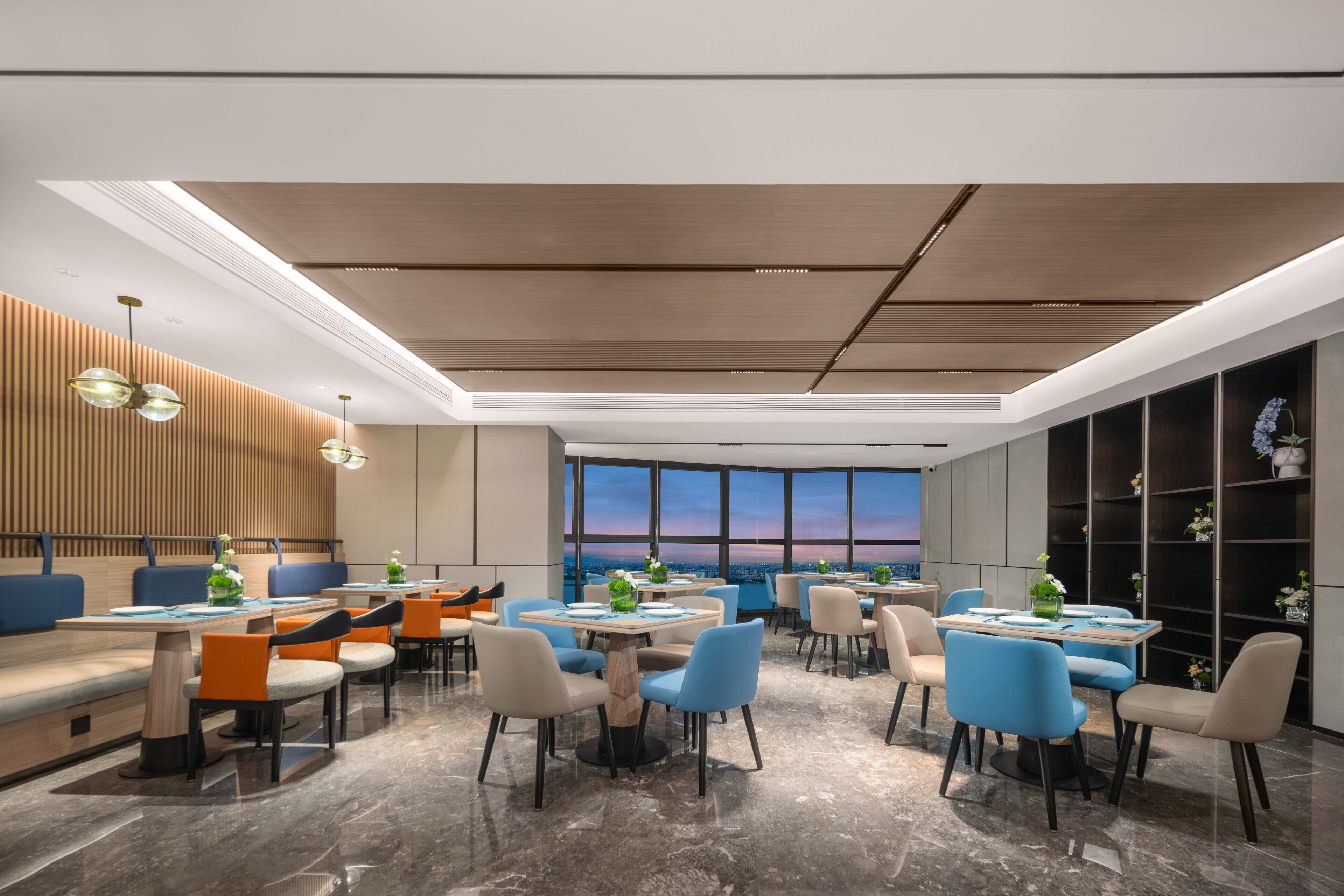 A modern dining area with multiple tables and chairs, large windows with a view, and a wooden ceiling.