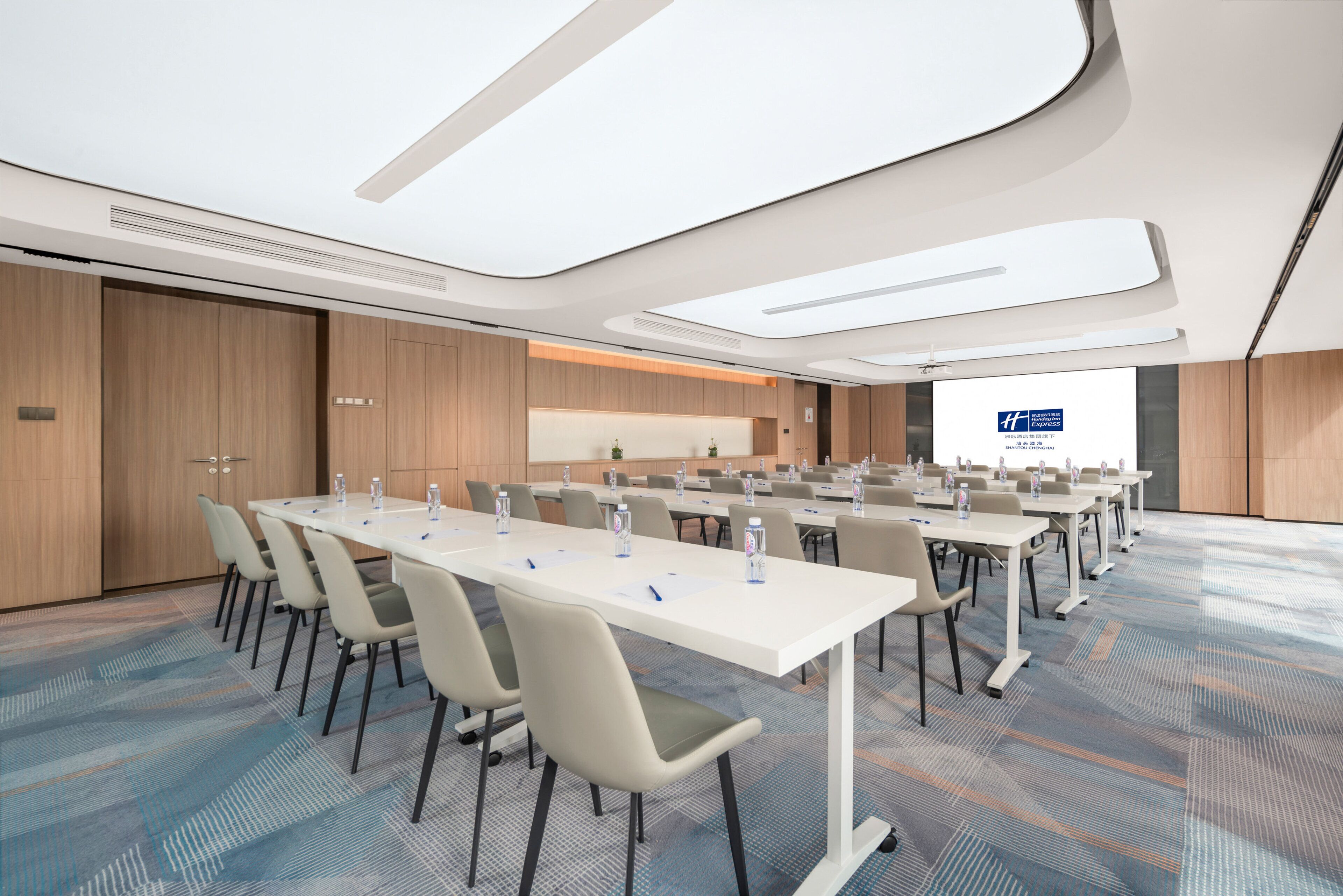 A conference room with long tables, chairs, and a large screen.