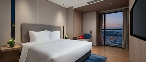 A modern hotel room with a large bed, a desk, a chair, and a large window offering a city view.