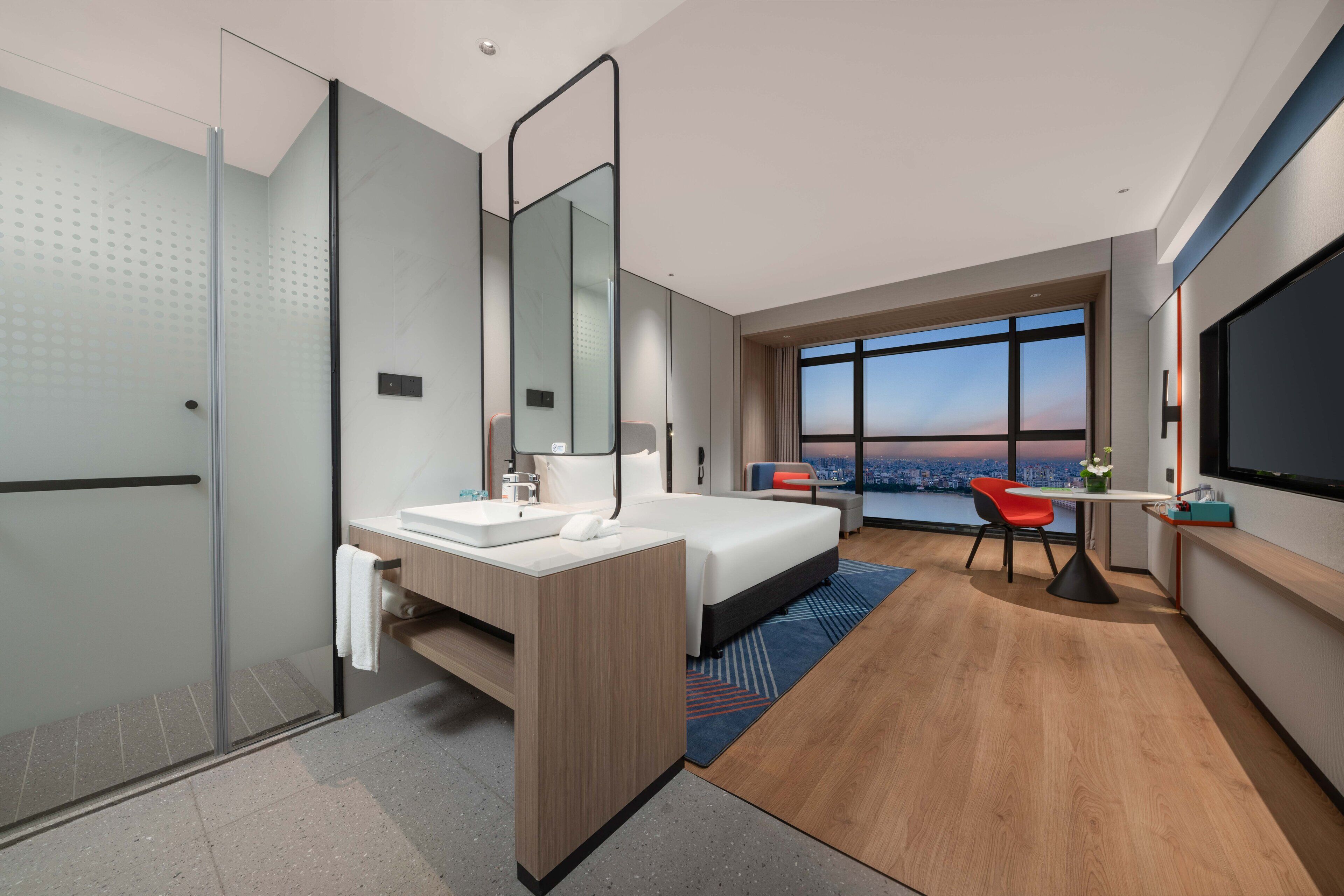 A modern hotel room with a large bed, a bathroom with a glass shower enclosure, a desk with a chair, and a large window offering a city view.