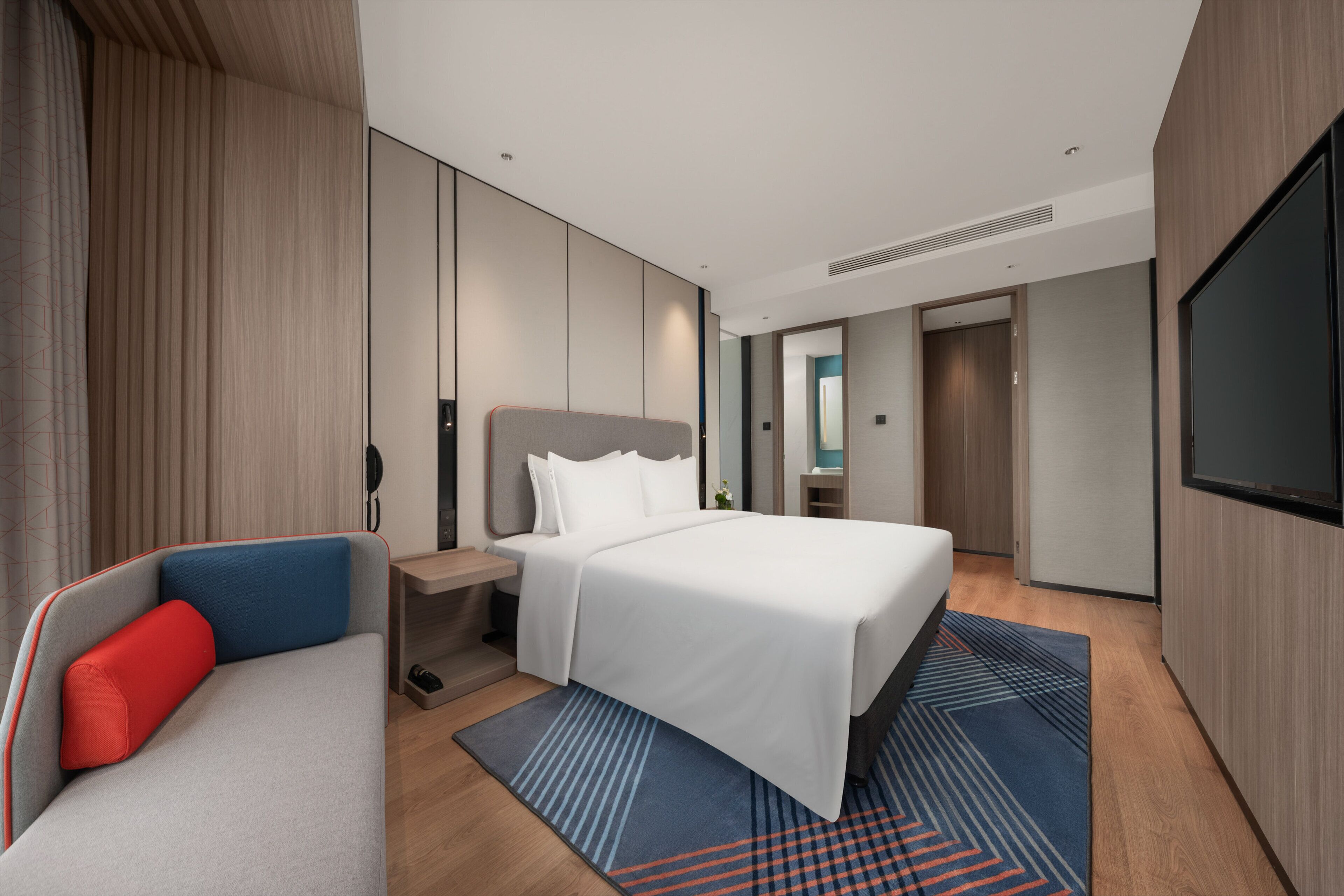 A modern hotel room with a bed, a seating area, a TV, and a bathroom visible through an open door.