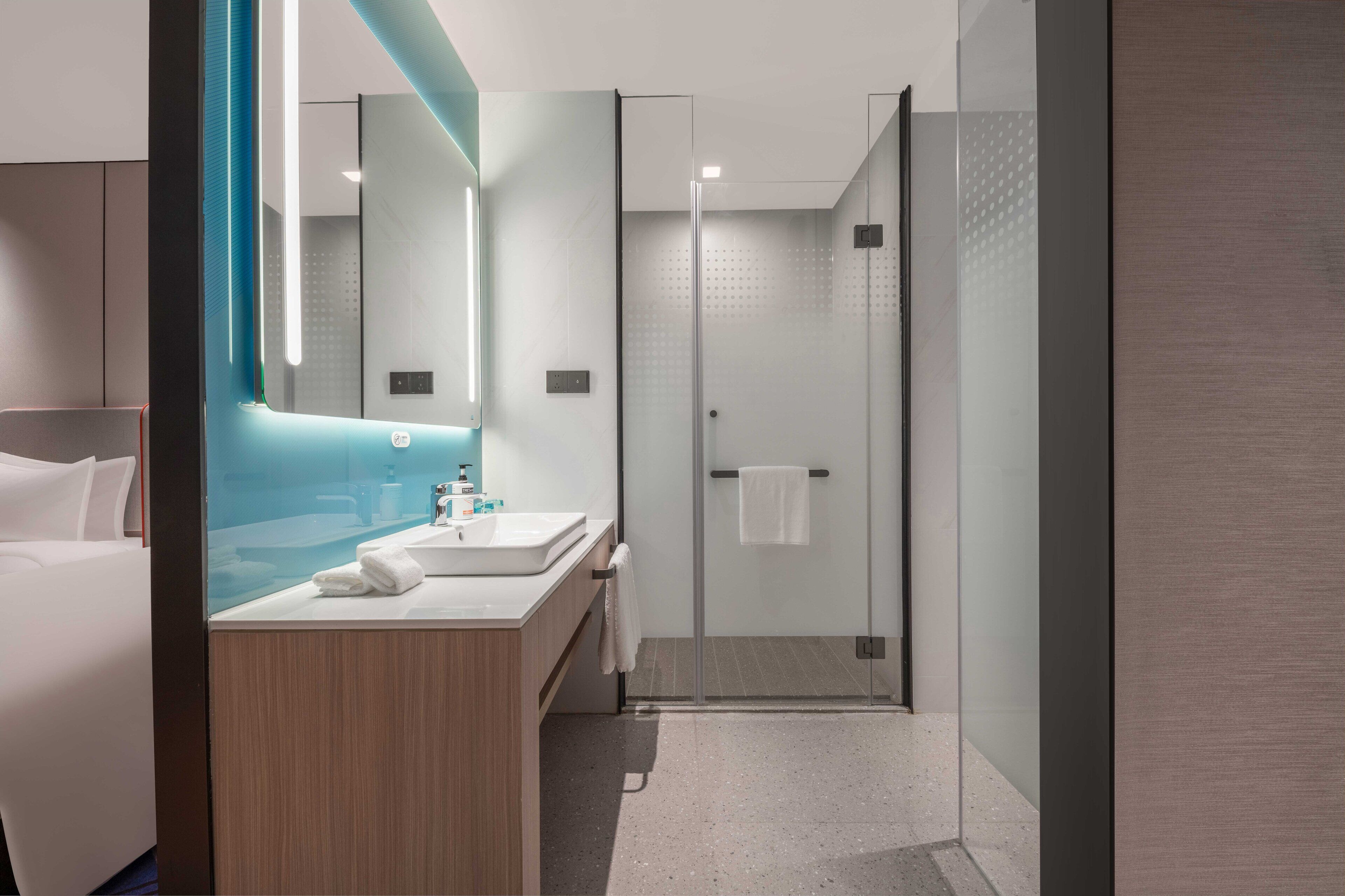 A modern hotel bathroom with a glass shower, a vanity with a mirror, and a bed in the adjacent room.