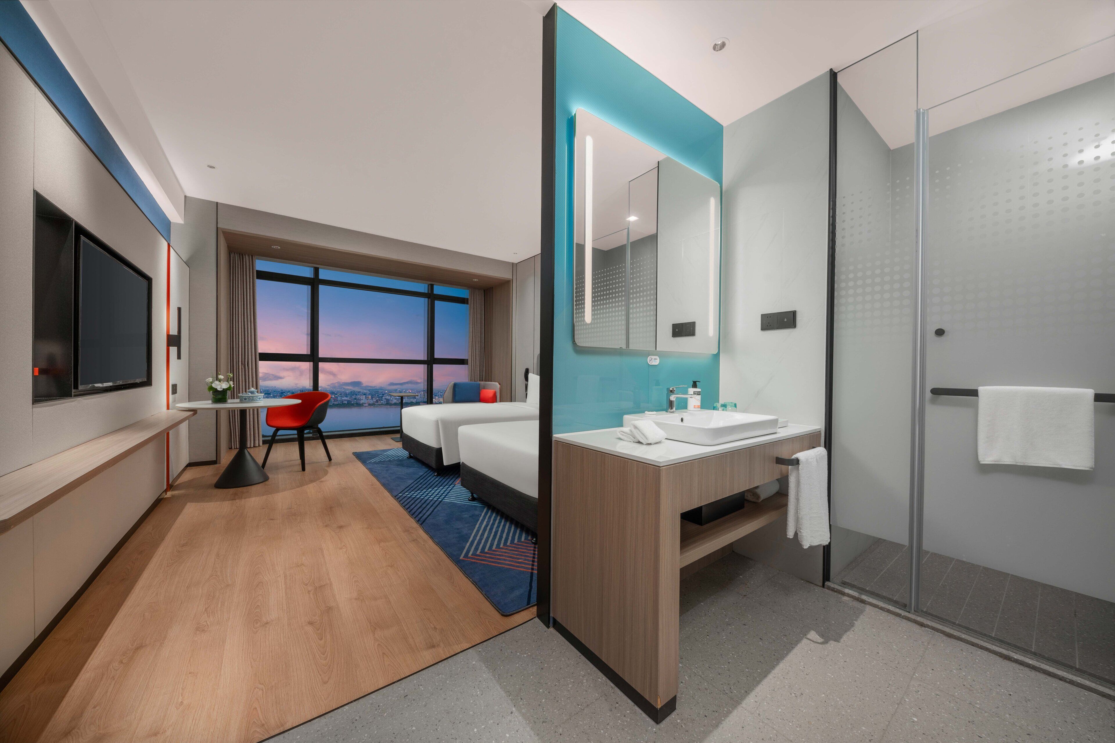 Una habitación de hotel moderna con un ventanal grande, una cama, un escritorio, una silla, un baño con lavamanos y espejo, y una zona de ducha.