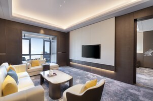 A modern living room with a large flat-screen TV mounted on the wall, a sofa set, a coffee table, and a large window offering a city view.