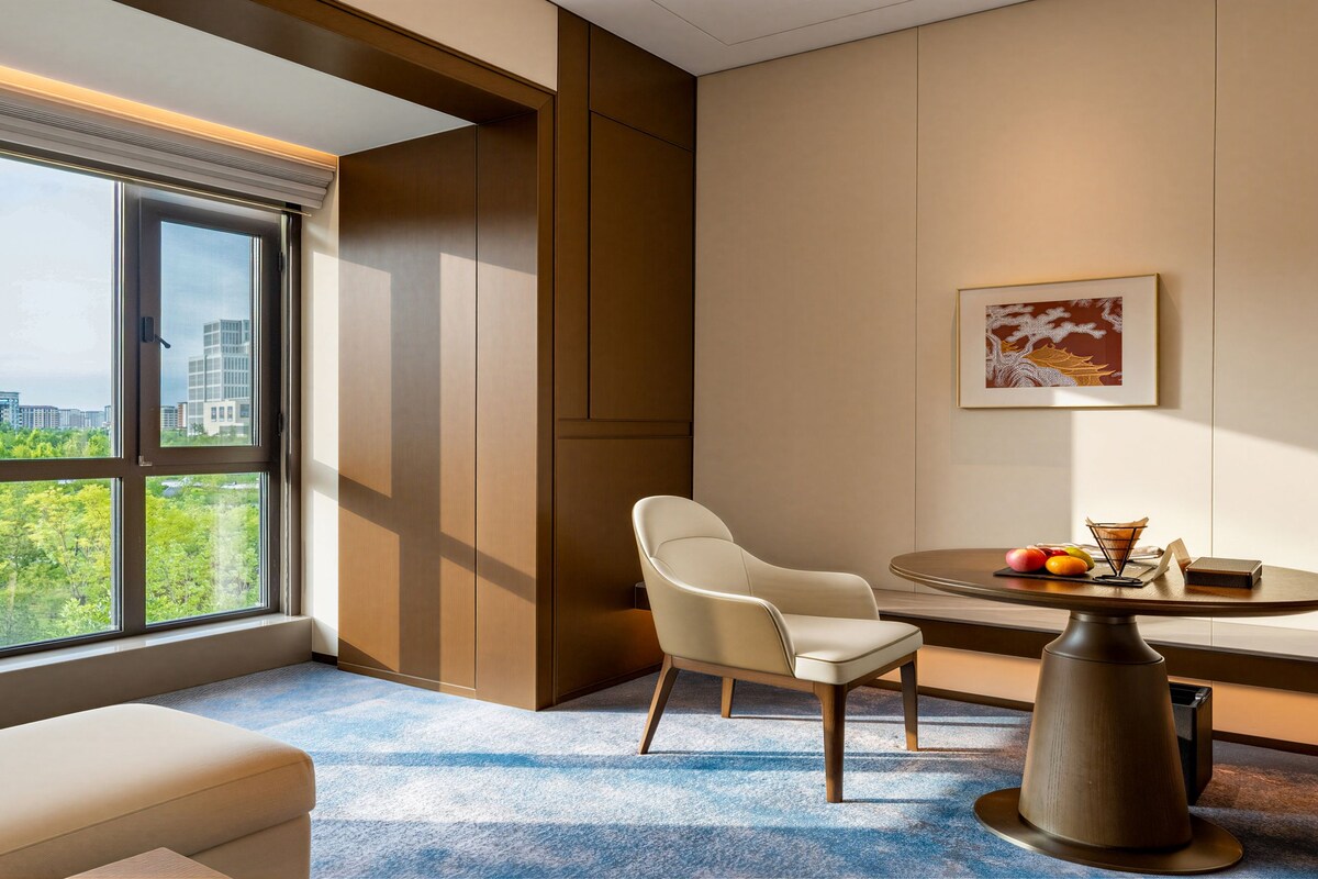 Habitación de hotel con un ventanal grande, una mesa redonda con una silla, un banco y un cuadro en la pared.