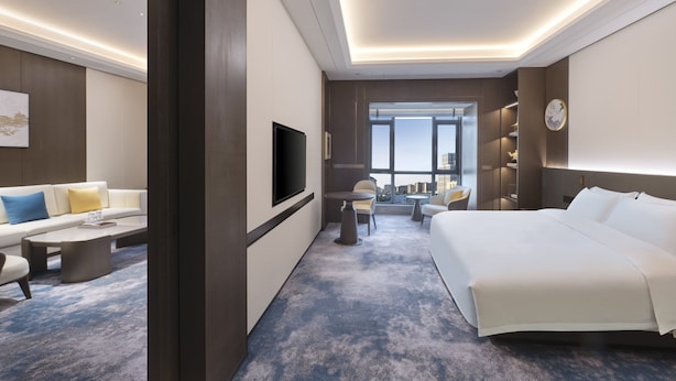 A modern hotel room with a large bed, a flat-screen TV, a sofa, a coffee table, and a view of the city through large windows. - voco Xiongan by IHG (Baoding)