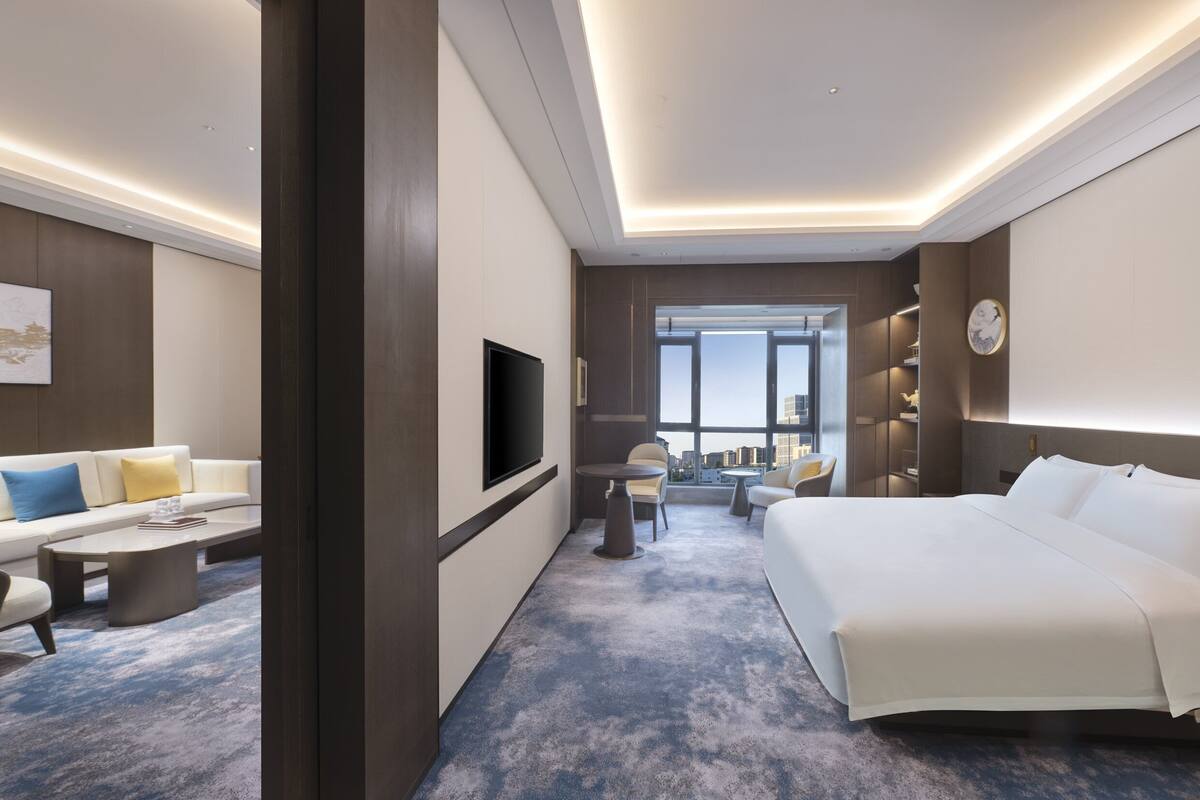 A modern hotel room with a large bed, a flat-screen TV, a sofa, a coffee table, and a view of the city through large windows.