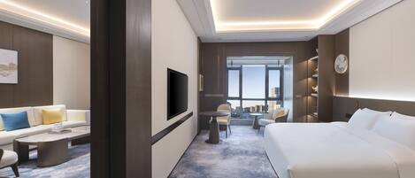 Una habitación de hotel moderna con una cama grande, un televisor de pantalla plana, un sofá, una mesita de café y vistas a la ciudad a través de amplios ventanales.