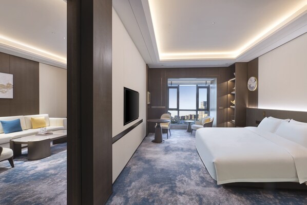 Quarto de hotel moderno com uma cama grande, televisão de tela plana, sofá, mesa de centro e vista da cidade por grandes janelas.
