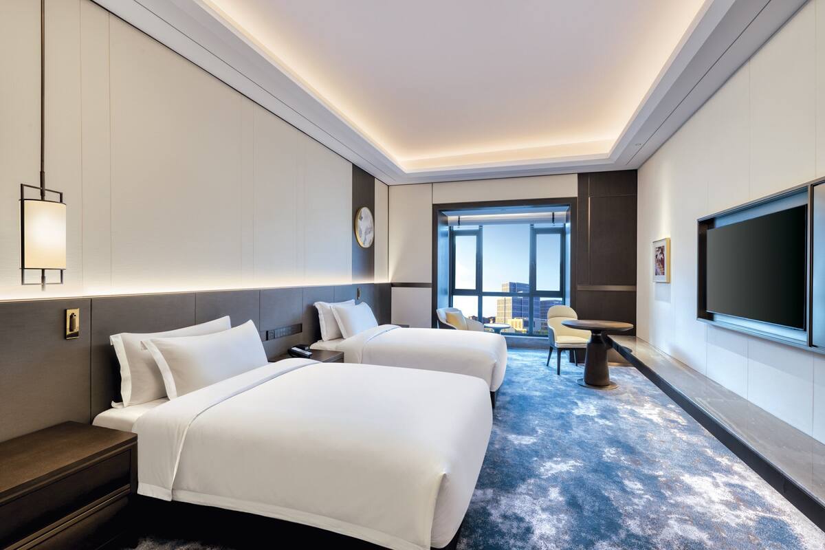 A hotel room with two beds, a desk, a chair, a TV, and a large window offering a city view.