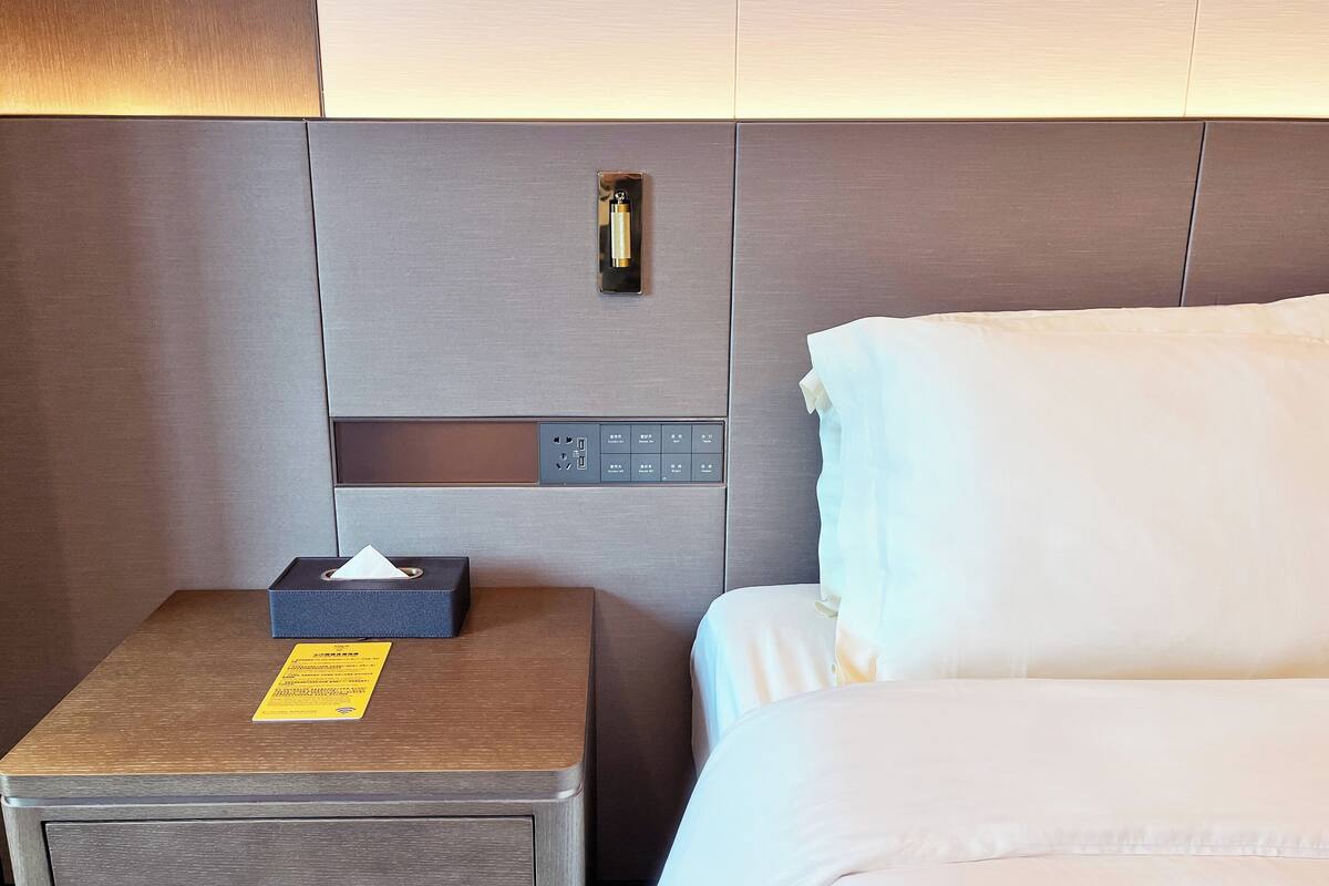 A hotel room with a wooden nightstand, a tissue box, a power outlet, and a control panel.