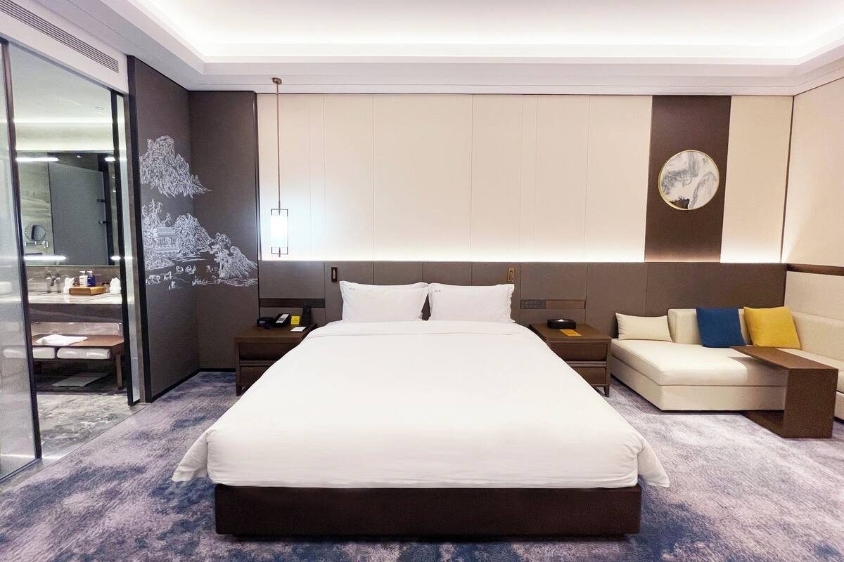 A modern hotel room with a large bed, a bedside table with a lamp, a sofa with cushions, and a large mirror.
