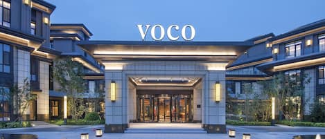 A modern hotel entrance with a prominent 'VOCO' sign, a covered entrance, and a landscaped area with illuminated planters.