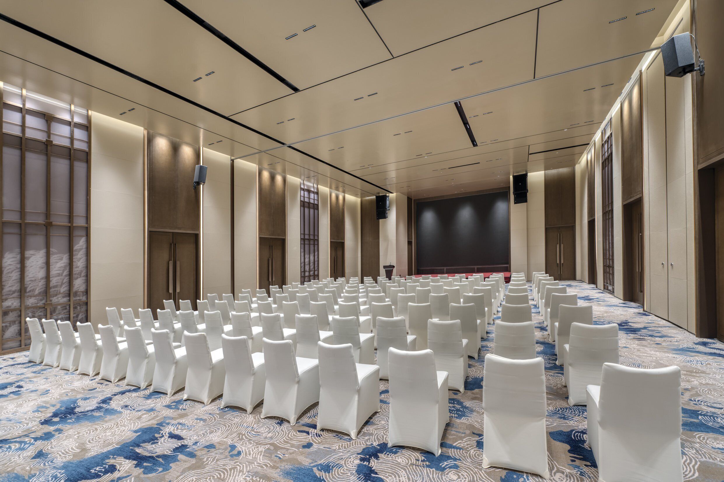 Een ruime congreszaal met rijen witte stoelen en een kamerbreed tapijt met dessin.